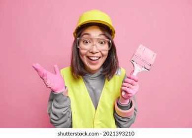 Positive Female Maintenance Worker Ready To Work Poses With Painting Brush Refurbishes Walls Of House Wears Uniform Hardhat Safety Glasses Isolated Over Pink Background. Decoration And Building