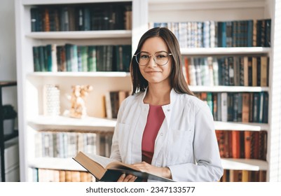 Positive female medical student in white uniform and eyeglasses standing near bookcase while reading book and looking at camera smiling - Powered by Shutterstock