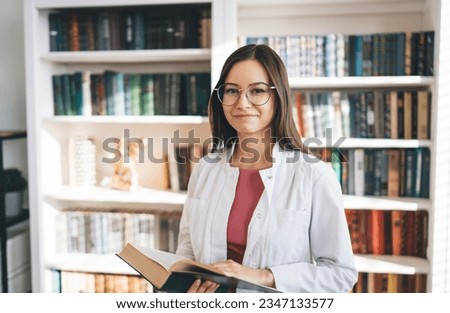 Positive female medical student in white uniform and eyeglasses standing near bookcase while reading book and looking at camera smiling