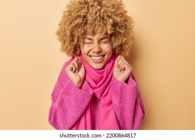 Positive Female Model With Curly Blonde Hair Keeps Eyes Closed Smiles Toothily Clenches Finsts Celebrates Good News Dressed In Pink Jumper And Scarf Around Neck Isolated Over Beige Background