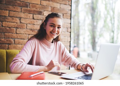 Positive Female Remote Worker In Casual Clothes With Toothy Smile Looking At Camera Sitting At Table With Opened Laptop And Doing Work Task