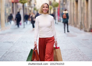 Positive female shopaholic carrying paper bags while strolling on pavement and looking at camera - Shutterstock ID 2212761313