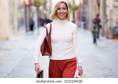 Positive female shopaholic with handbag carrying paper bags with purchases after shopping in stores and smiling at camera - Shutterstock ID 2122139630