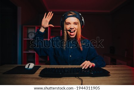 Positive female streamer sits at night in the room at the computer and communicates on a headset with subscribers, poses palms of greetings to the camera and smiles.