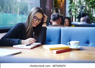 Positive female student in stylish eyeglasses with good lense writing down funny story in notebook sitting in coffee shop with cup of cappuccino and books.Smiling female noting text in notepad