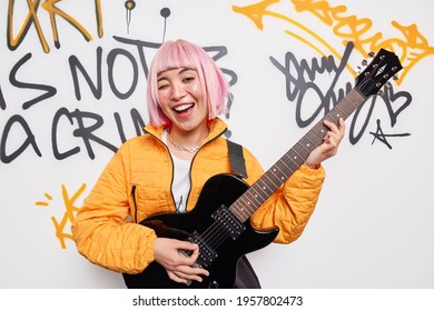 Positive female teenage rocker plays electric guitar has fun entertains friends at street has trendy pink hair wears orange jacket poses against graffiti wall. Happy guitarist plays favorite music