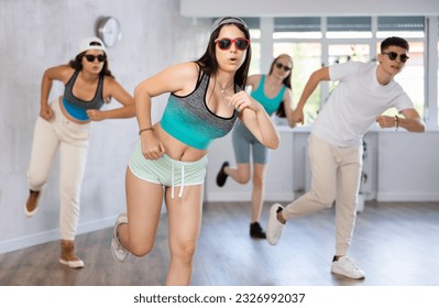 Positive female teenager dances shuffle in choreographic school, group of young people in sportswear and dark glasses ball cap trains together with their classmates in gym before competitions, battle