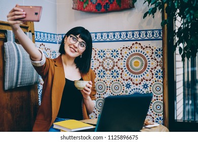 Positive Female Teenager Enjoying Tea Time Taking Images On Front Mobile Camera, Cheerful Woman Sitting Near Bohemian Wall And Making Video For Blog Using Multimedia Application On Cellular Phone