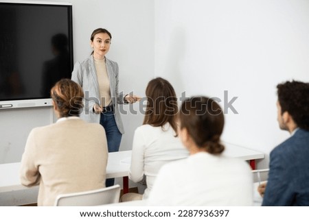 Positive female trainer standing near interactive board and conducting advanced training courses to office employees sitting at desks in classroom