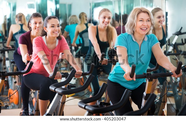 Positive females of different age\
training on exercise bikes together in modern fitness club\
