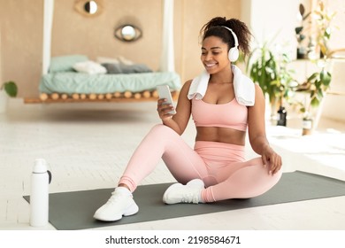 Positive Fit Black Woman With Towel On Shoulders And Wireless Headphones Sitting On Fitness Mat And Listening To Music While Resting During Home Workout, Free Copy Space