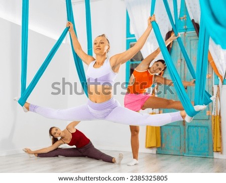 Positive fit woman practicing antigravity yoga with group in fitness studio, doing aerial split, placing feet on suspended hammocks