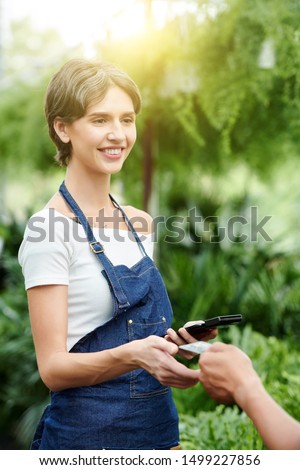 Positive florist in denim apron taking credit card from hand of customer to accept payment