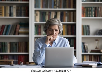 Positive focused senior business lady using laptop at table in home office, library with bookshelves, watching learning webinar, online video presentation, thinking over project, work tasks - Shutterstock ID 2156185777