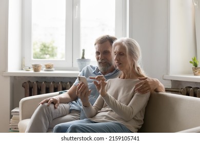Positive focused senior couple using online app on smartphone, internet virtual payment service on mobile phone, making video call, shopping on we stores, resting on couch at home