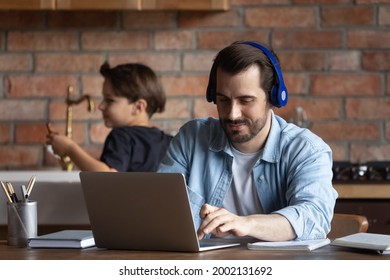 Positive Freelance Man In Headphones Working At Laptop From Home With Little Kid In Background. Adult Student Using Online App, Watching Learning Webinar, Video Lesson, Attending Virtual Class,