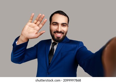 Positive Friendly Bearded Businessman Making Selfie Or Streaming, Waving Hand POV, Point Of View Of Photo, Wearing Official Style Suit. Indoor Studio Shot Isolated On Gray Background.