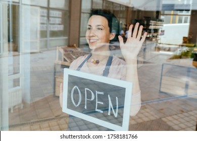 Positive Friendly Charming French Business Woman Face Against Smiling Waitress Or Owner Showing Chalkboard With Open Sign Through Window. Quarantine, Coronavirus, Isolation. Back To Working Life..