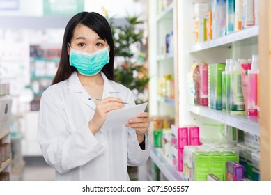 Positive Friendly Smiling Chinese Female Pharmacist In Protective Facial Mask Standing Among Shelves In Drug Store