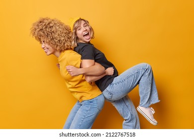 Positive friends give piggyback ride for each other foolish around dressed in casual clothes laugh joyfully isolated over yellow background feel lively and energetic. Two wmen have fun indoor