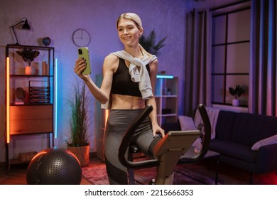 Positive funny blond caucasian sports women in sportswear doing selfie on her mobile phone, cycling bike at home at night time. Cardio training, exercising legs, cardio workout indoors. - Powered by Shutterstock