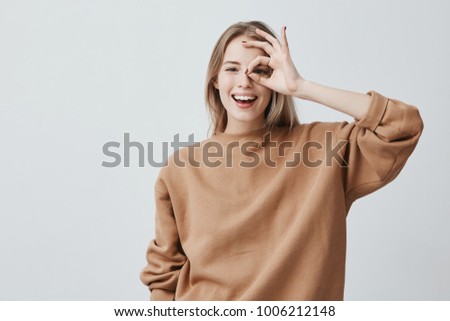 Positive funny blonde girl in casual clothes shows ok sign, laughs at camera, demonstrates that everything is fine, agrees. Cheerful woman gestures indoors. Body language and human emotions