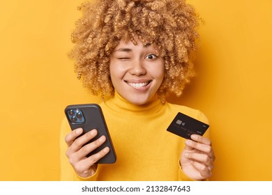 Positive funny curly haired woman winks eye smiles toothily thinks of online shopping holds mobile phone and banking card makes payment dressed in casual jumper isolated over yellow background. - Powered by Shutterstock