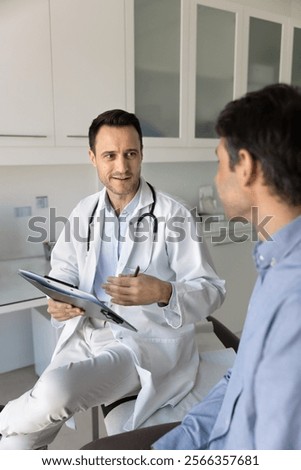 Positive general practitioner man talking to patient on appointment in clinic office, holding pad with medical records, speaking, discussing complaints, recommendation, explaining treatment