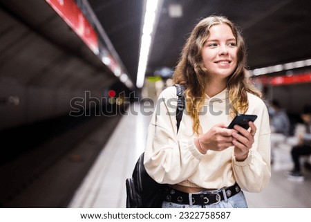 Positive girl with a backpack standing on the platform of a metro station, texting on her mobile phone with friends, ..waiting for the train