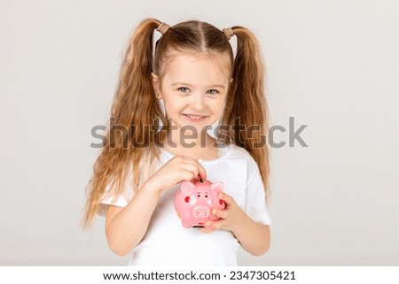 Positive girl in casual outfit laughing and saving money in piggy bank against white background