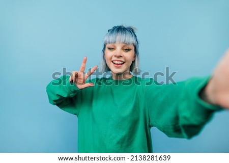Positive girl in a green sweater and blue hair takes a selfie with a smile on his face and shows a gesture of peace isolated on a blue background