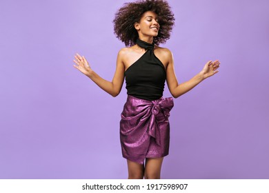 Positive girl in purple skirt jumping on lilac backdrop. Wavy haired woman in black top smiles with closed eyes on isolated background.. - Shutterstock ID 1917598907