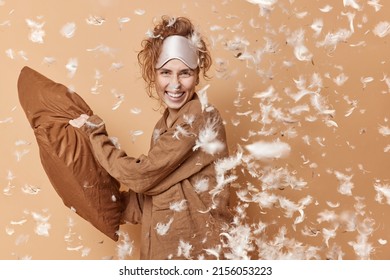 Positive glad young woman has pillow fight after awakening has fun and laughs dressed in comfortable pajama blindfold on forehead poses indoor feathers flying around. Sleeping rest home concept