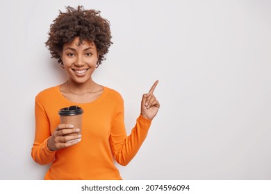 Positive good looking woman with curly hair wears casual orange jumper drinks takeaway coffee points at upper right corner shows copy space for your advertising content. Look at this please. - Powered by Shutterstock