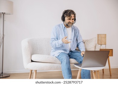 Positive grandfather using laptop computer and headset, sitting on couch. Senior man working from home, have online meeting, video chat, copy space - Powered by Shutterstock