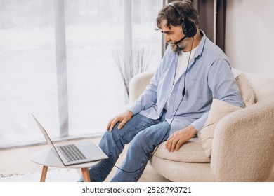 Positive grandfather using laptop computer and headset, sitting on couch. Senior man working from home, have online meeting, video chat, copy space - Powered by Shutterstock