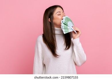 Positive Greedy Girl With Dark Hair Holding Big Fan Of Euro Banknotes, Smelling Money, Lottery Winner, Wearing White Casual Style Sweater. Indoor Studio Shot Isolated On Pink Background.