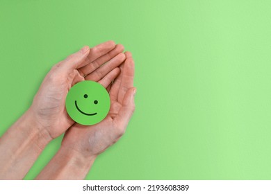 A Positive Green Smiley Face Being Held In A Woman's Hands Against A Cheerful Green Paper Background.