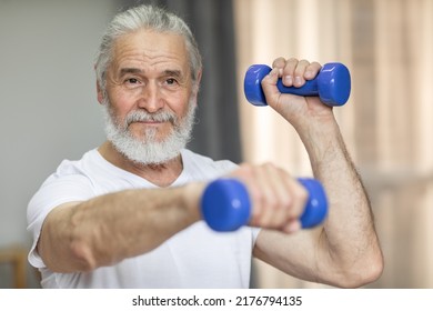 Positive grey-haired bearded old man in sportswear exercising alone at home, using blue barbells, working on arms strength, pumping iron and smiling at camera, closeup photo - Powered by Shutterstock