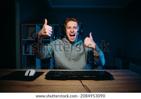Positive guy in a headset plays computer games and rushes with a serious face on the background of a cozy room at night.