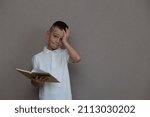 Positive haired elementary school nerd boy  holding open textbook and looking at camera against the wall