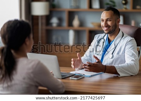 Positive handsome arab man doctor consulting unrecognizable african american lady patient, doc giving recommendations, smiling and gesturing, sitting at workdesk with laptop at clinic