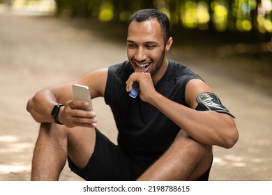 Positive Handsome Black Guy Athlete Having Snack, Resting After Workout Outdoors In Public Park, Sitting On The Ground, Eating Protein Bar And Using Smartphone, Checking Nice Fitness Mobile App