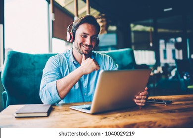 Positive Handsome Businessman In Modern Headphones Watching Funny Webinar About Productive Work Strategy On Modern Digital Computer Connected To 4G Internet Sitting In Stylish Cafeteria