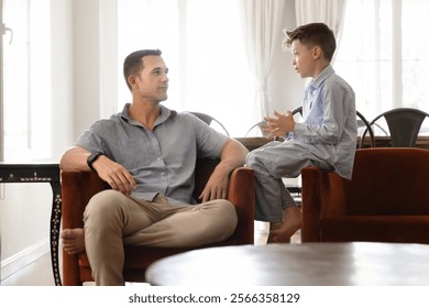 Positive handsome dad talking to son kid at home, sitting in cozy armchair, listening to boy with interest, involvement, giving advice, enjoying family relationship, fatherhood
