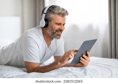 Positive handsome grey-haired bearded middle aged man in homewear lying on bed at home, using modern wireless headphones and digital tablet, touch screen, choosing video content online, free space - Powered by Shutterstock