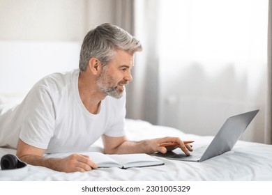 Positive handsome grey-haired middle aged man wearing pajamas lying on bed at home, using modern pc laptop, taking notes in notepad, attending online training or webinar, e-education