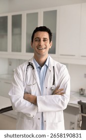 Positive handsome Hispanic general practitioner man in white coat and stethoscope standing with hands folded in hospital exam room, looking at camera for head shot, smiling. Vertical shot