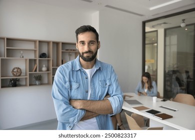 Positive handsome Hispanic project leader man posing in office meeting room space, looking at camera with arms crossed, standing for professional portrait with colleagues working in background - Powered by Shutterstock