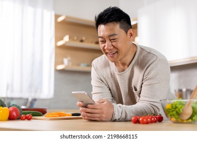 Positive handsome middle aged asian man single father holding cell phone, checking newest mobile application to learn cooking, kitchen interior, copy space. Free recipe mobile app, culinary app - Powered by Shutterstock